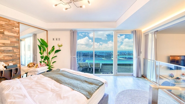 Une chambre moderne avec un grand lit, une vue sur une piscine et sur le paysage urbain grâce à de grandes fenêtres du sol au plafond, et un balcon doté d’une rambarde en verre.