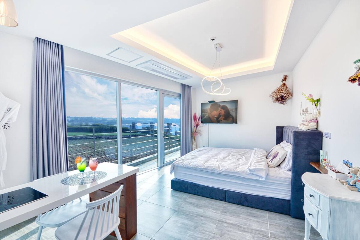 Ein modernes Hotelzimmer mit einem großen Balkon, einem Bett mit blauer Kopfteil, einem Schreibtisch mit Stuhl und Blick auf die Stadtlandschaft.