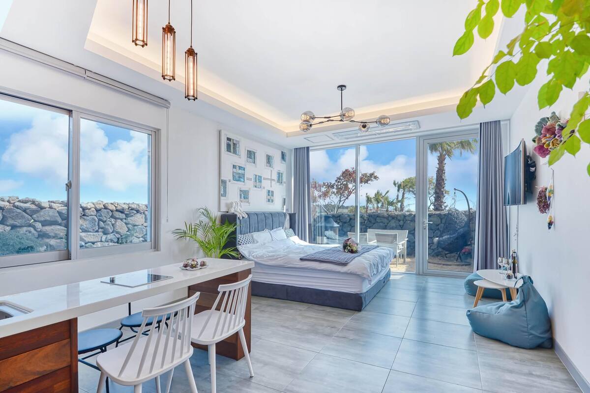 Ein modernes Schlafzimmer mit einem großen Bett, einem Schreibtisch und Stühlen, einem Balkon mit Blick auf Himmel und Landschaft sowie einem an der Wand montierten Fernseher.