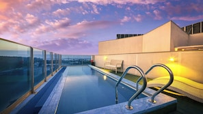 A rooftop infinity pool with glass railings, a bench, and a view of the cityscape.