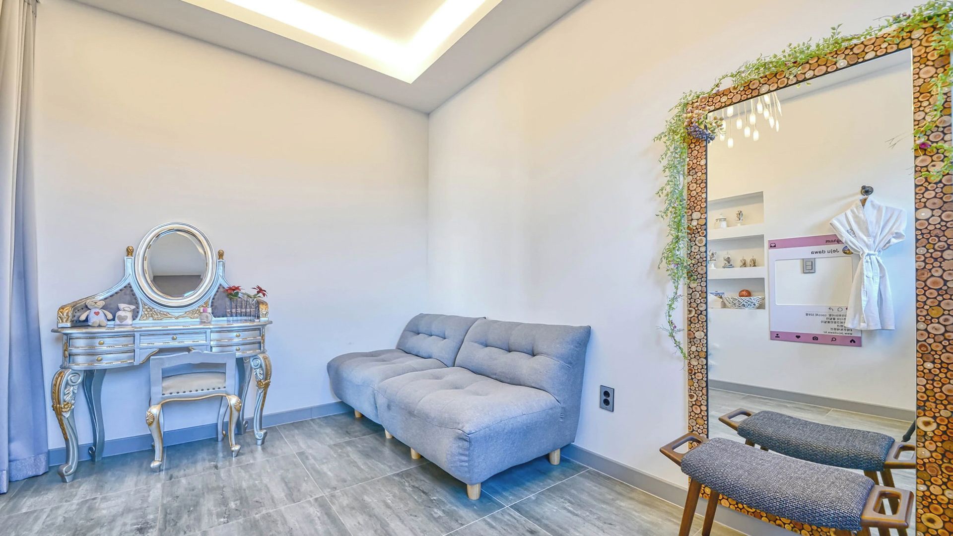 Une pièce comprenant une coiffeuse, un canapé gris, un miroir avec un cadre décoratif et une chaise.