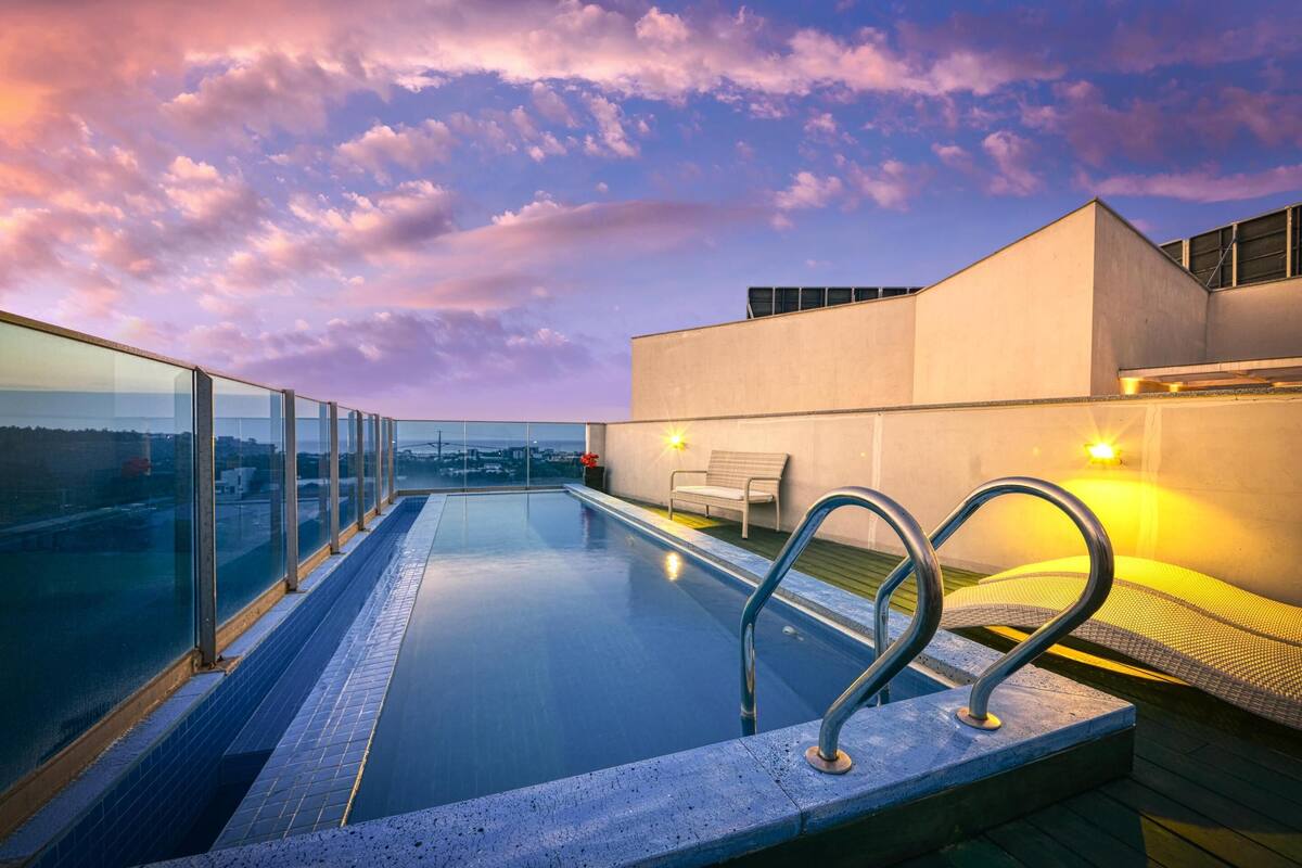 Ein Pool mit Infinity-Kante auf einem Dach, mit Glasgeländern, einer Bank und einem modernen Gebäude im Hintergrund.