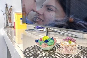 Un televisor mostrando un primer plano del rostro de una persona, un cuenco decorativo con objetos coloridos y un pastel pequeño sobre una mesa.