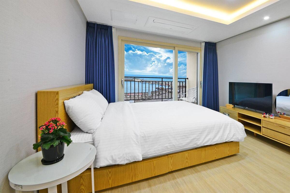 Habitación de hotel con una cama grande, un televisor, una mesita de noche con una planta y vista al mar desde el balcón.