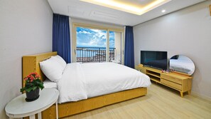 Une chambre d’hôtel avec un grand lit, une télévision, une table de chevet agrémentée d’une plante, et une vue sur la mer depuis le balcon.