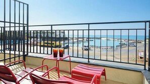 Un balcon avec des chaises rouges et une table, donnant sur une marina où sont amarrés des bateaux, et sur une plage.