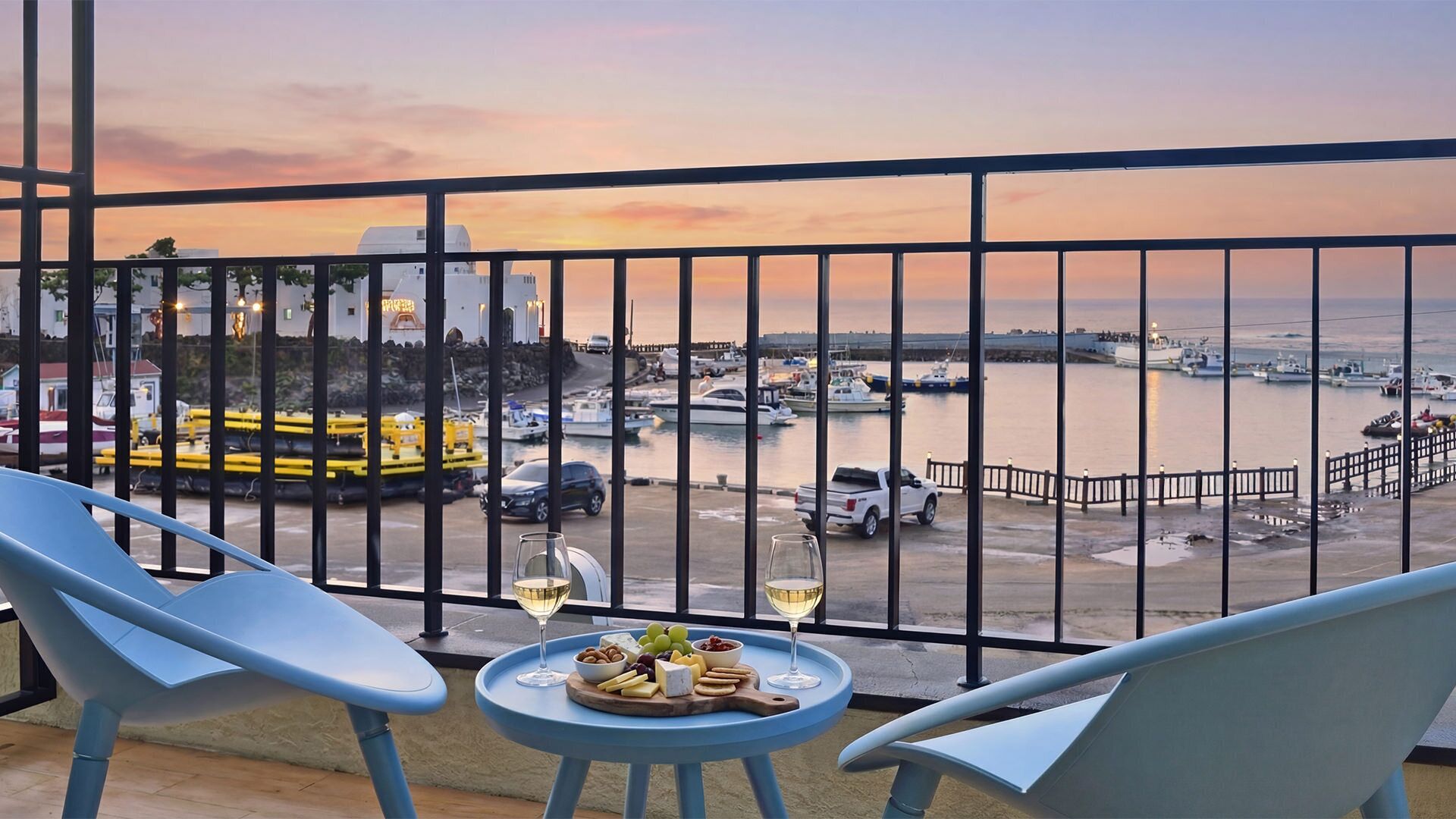 A balcony with two chairs, a small table with a cheese platter and two glasses of wine, overlooking a marina with boats and a sunset.