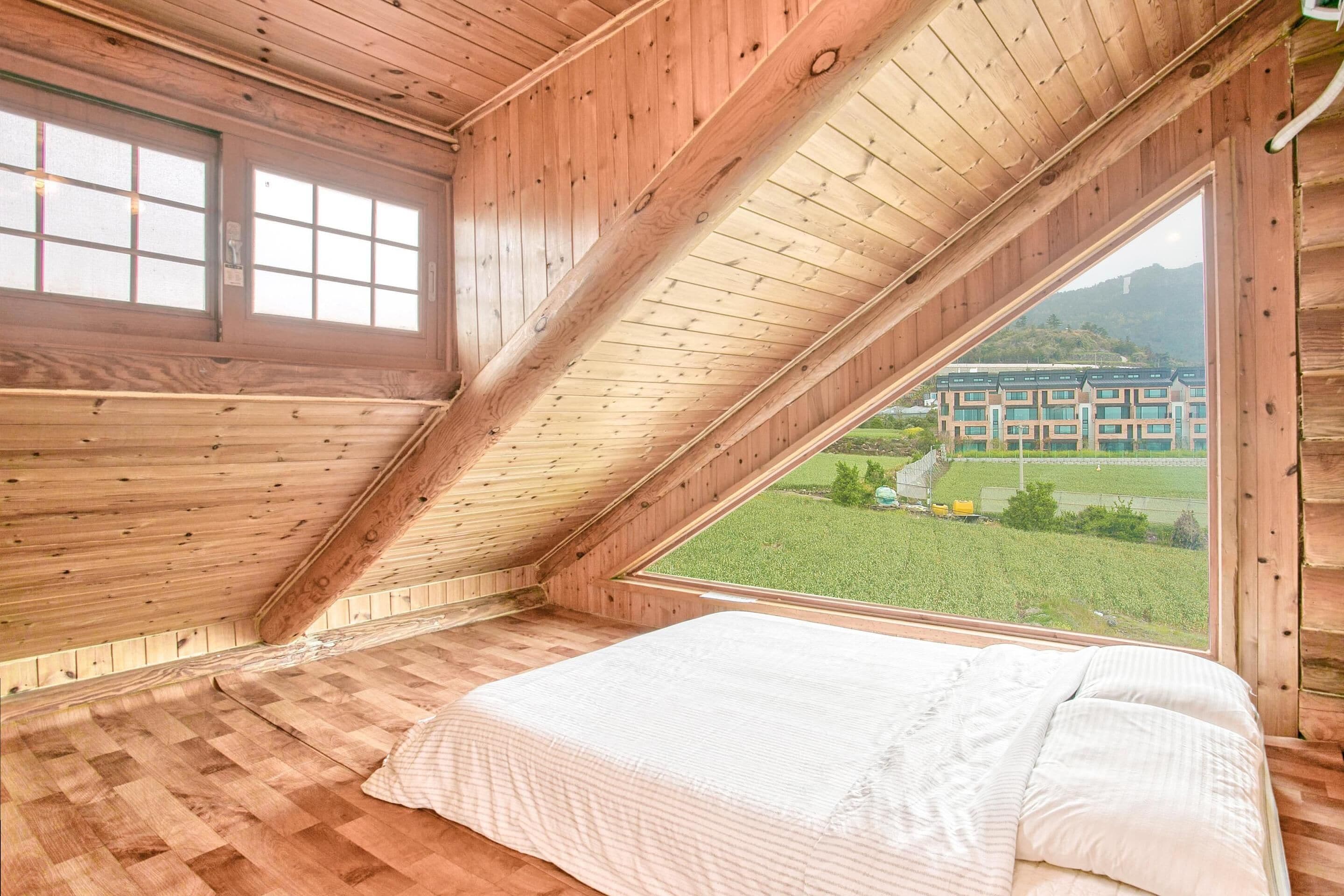 A room with a sloped ceiling, wooden walls, and a large window offering a view of a building and greenery.