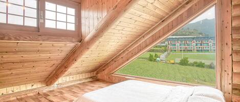 A room with a sloped ceiling, wooden walls, and a large window offering a view of a building and greenery.