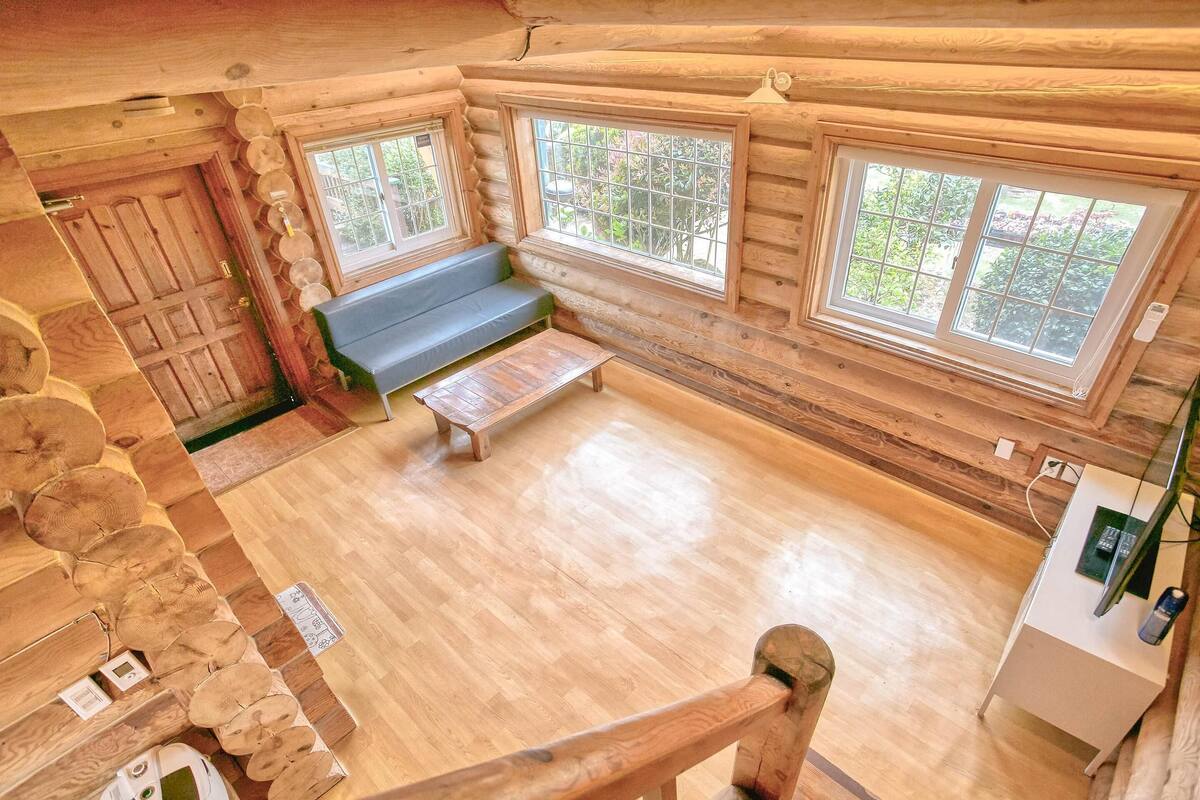 A log cabin interior with a wooden floor, a bench, and two windows offering a view of greenery.