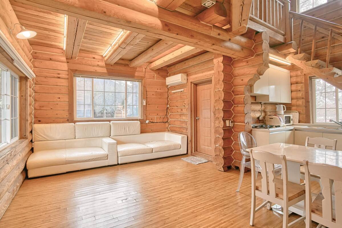 A log cabin interior with a wooden floor, a white sofa, a dining table with chairs, and a kitchen area.
