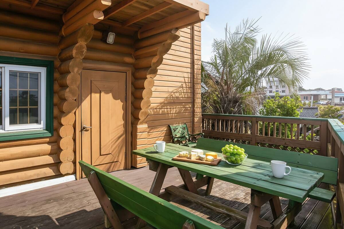 A wooden cabin with a green-framed window, a door, and a deck with a table and chairs.