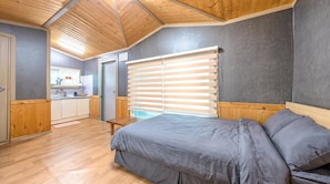 A bedroom with a bed, a window with blinds, a wooden ceiling, and a kitchenette in the background.
