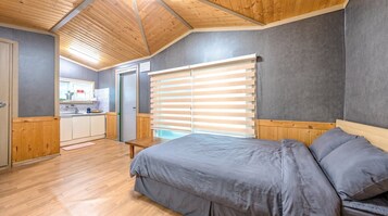 A bedroom with a bed, a window with blinds, a wooden ceiling, and a kitchenette in the background.