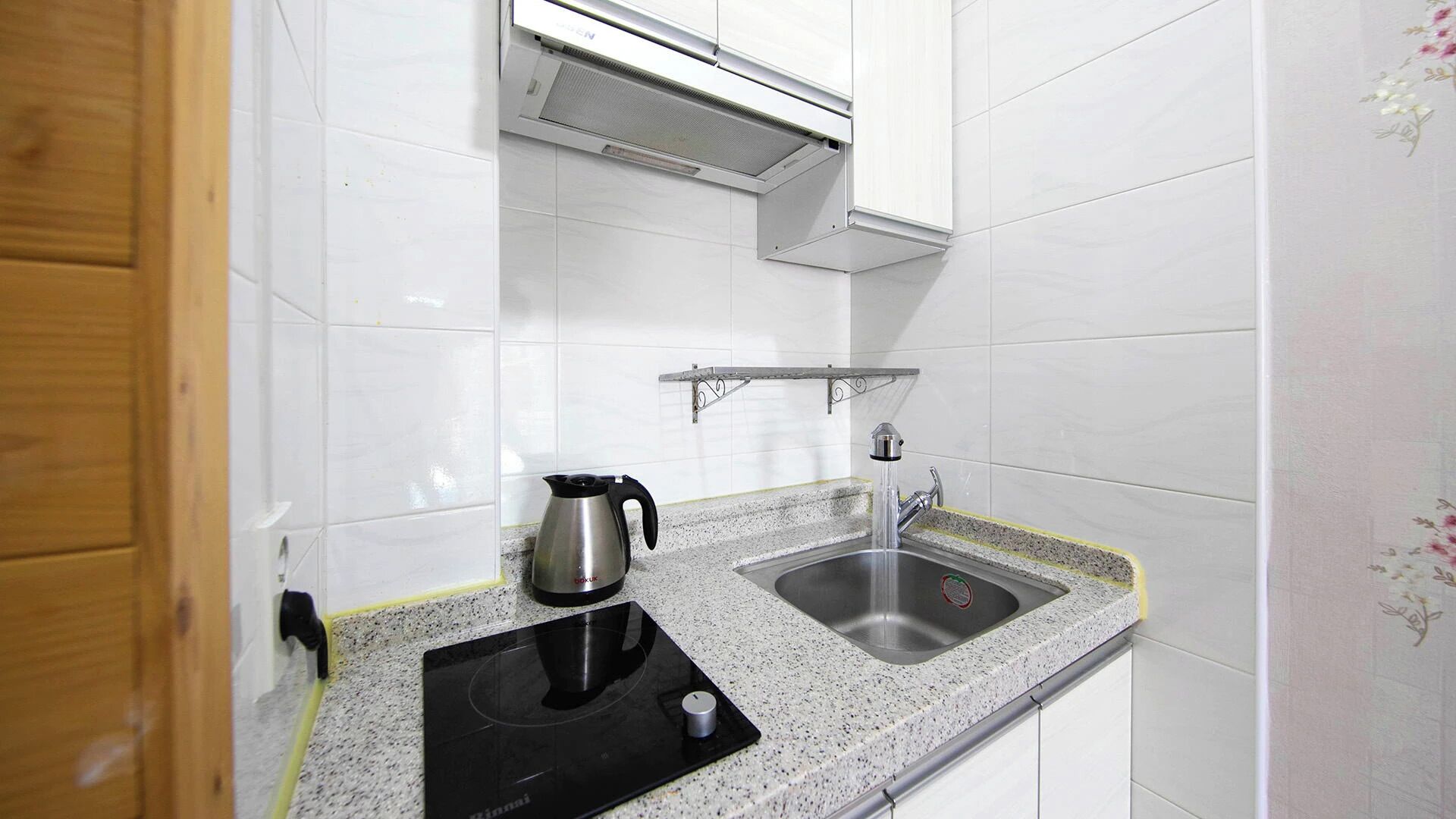 Une cuisine avec des carreaux blancs, un évier en acier inoxydable, une bouilloire et une plaque de cuisson à induction noire.