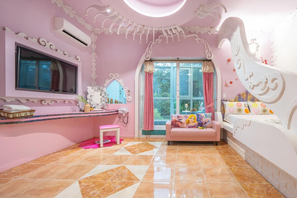 Una habitación con decoración rosa, un ventanal grande, un tocador con espejo y una cama con almohadones coloridos.
