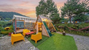 Ein Wintergarten mit gelben Stühlen und einem Esstisch, umgeben von einem Garten mit einer Hängematte und einem kleinen Teich.