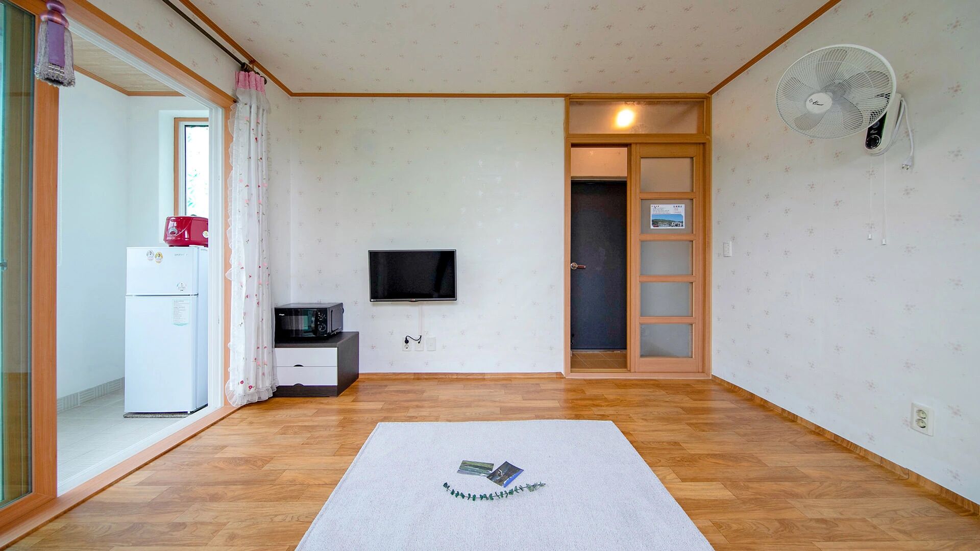 A room with a wooden floor, a flat-screen TV mounted on the wall, a white refrigerator, a microwave, and a fan.
