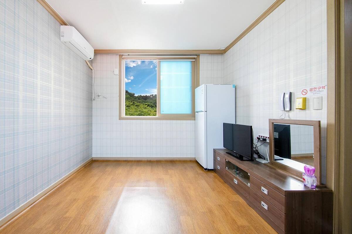 Una habitación pequeña y ordenada con piso de madera, una ventana que da a vistas de vegetación, un refrigerador, un televisor y un armario de madera.