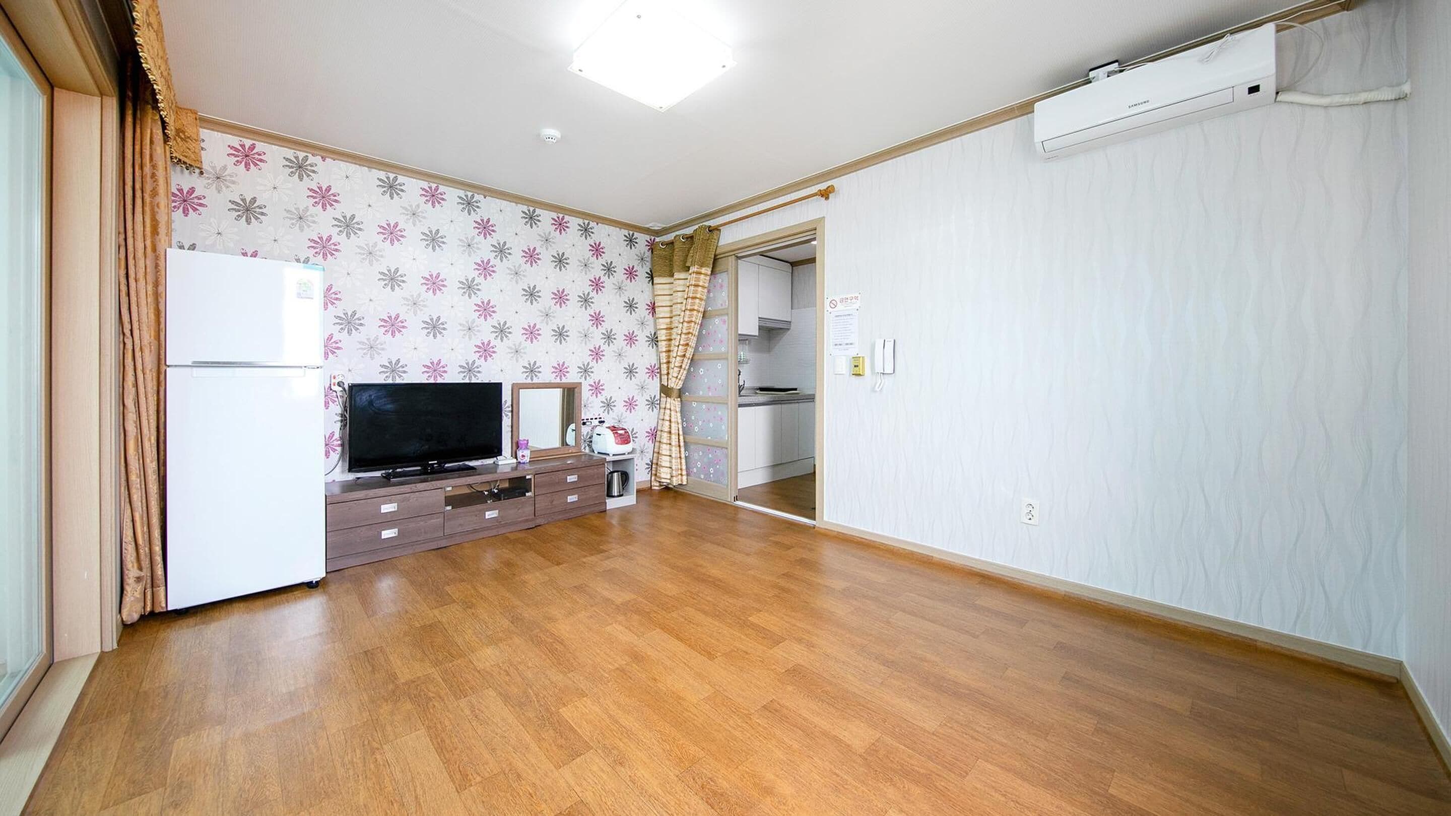 A spacious living room with a wooden floor, a flat-screen TV, a small kitchen area, and an air conditioning unit.