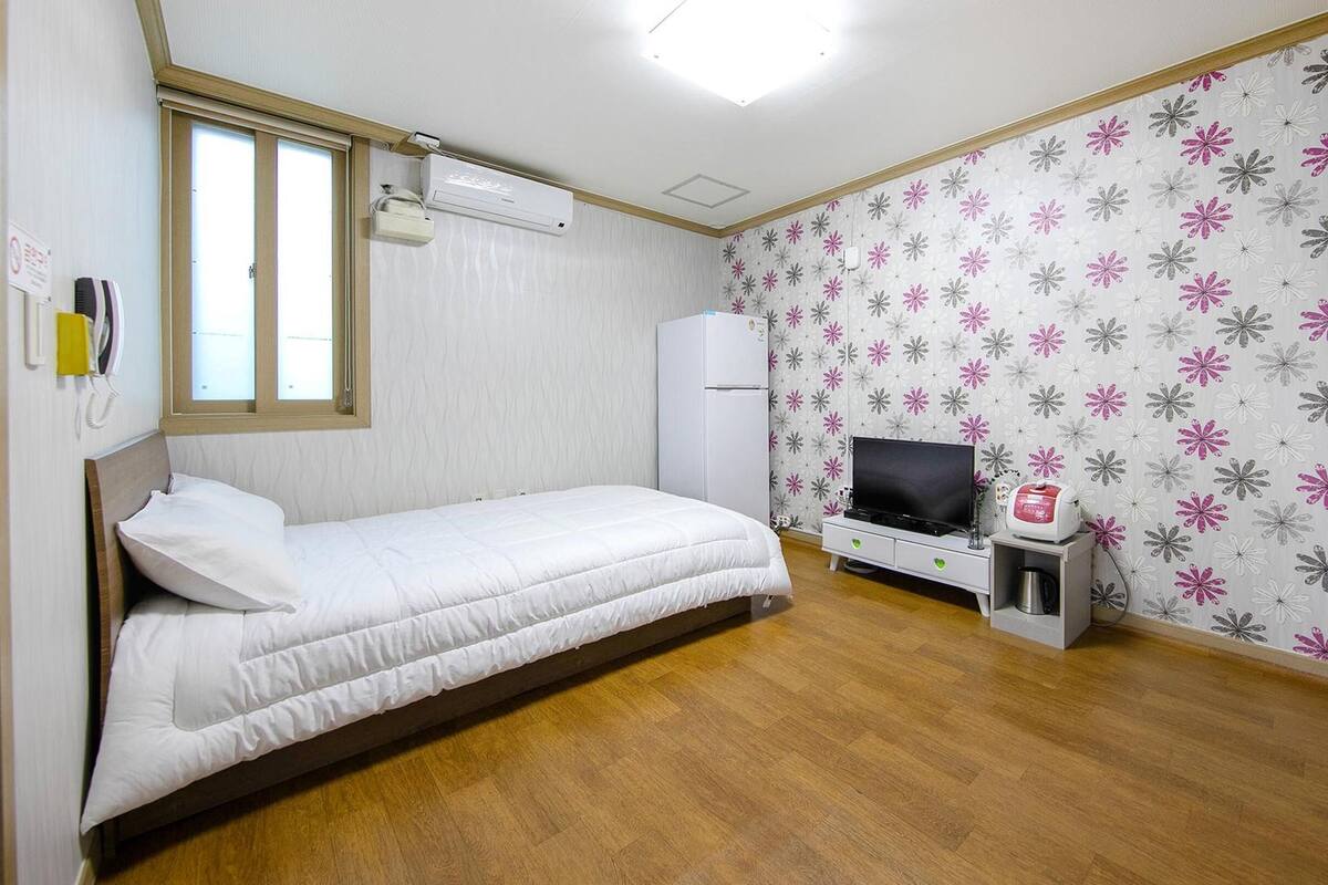 Una habitación pequeña con cama individual, piso de madera, cama blanca, televisor, mueble pequeño y refrigerador.