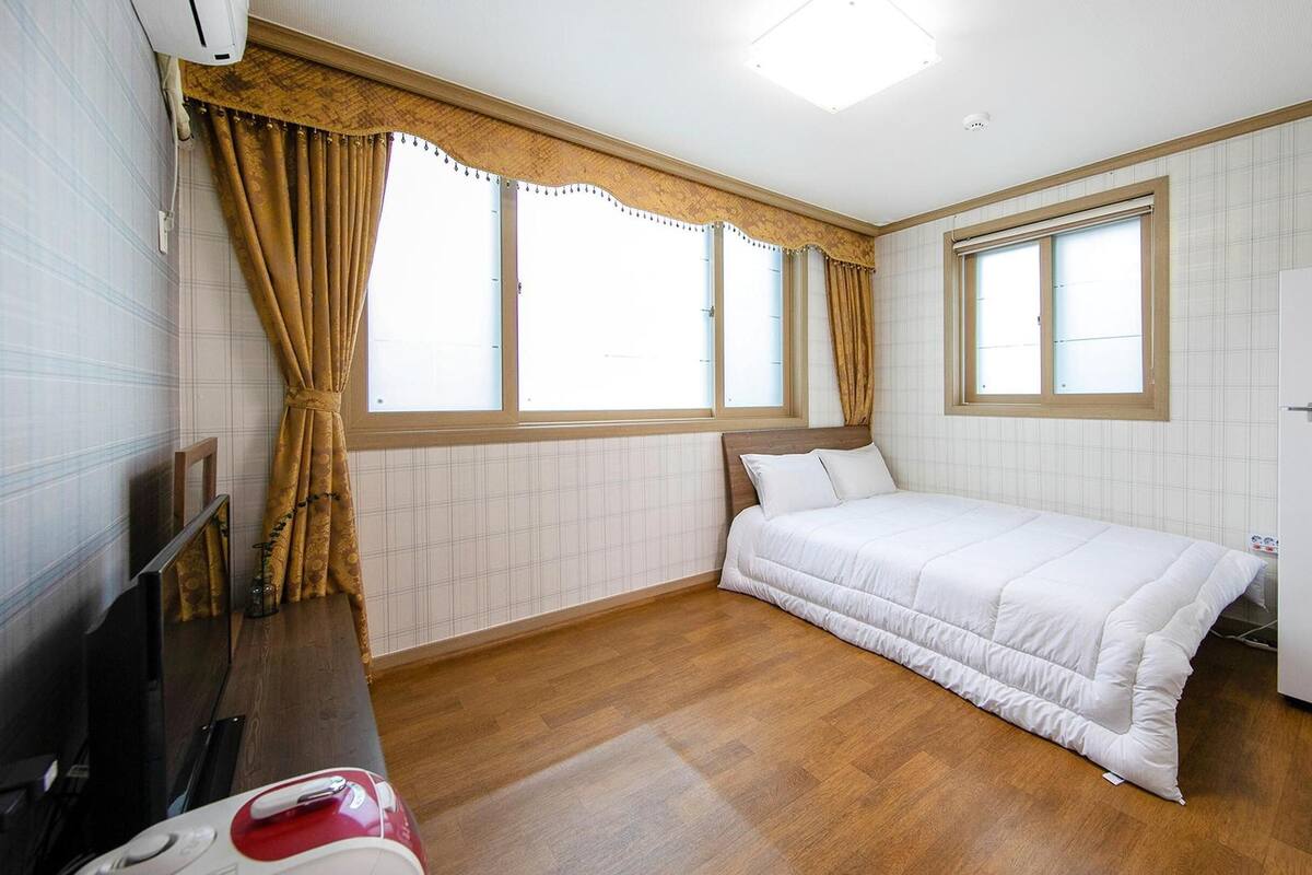 Una habitación pequeña con cama, refrigerador, ventana y televisor.