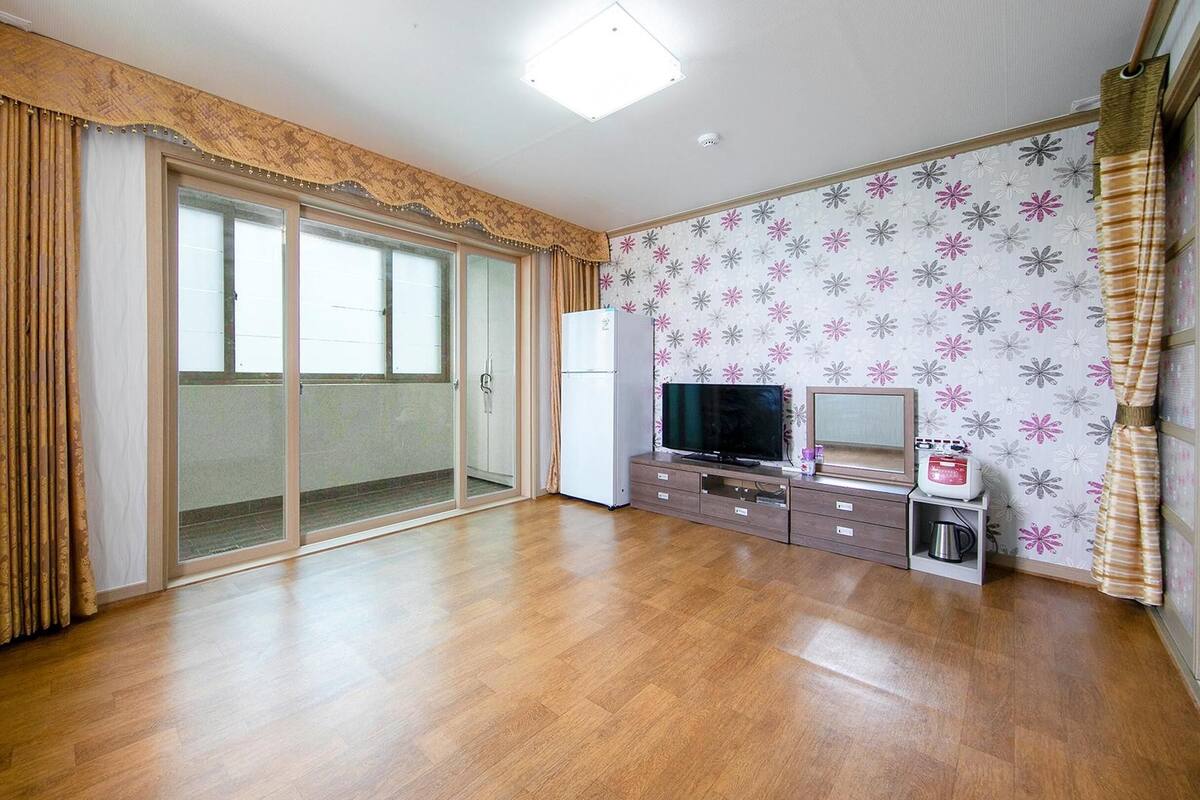 Una habitación con puerta de vidrio corredera que da a un balcón, un televisor de pantalla plana, un refrigerador pequeño y un microondas sobre un mueble de madera.