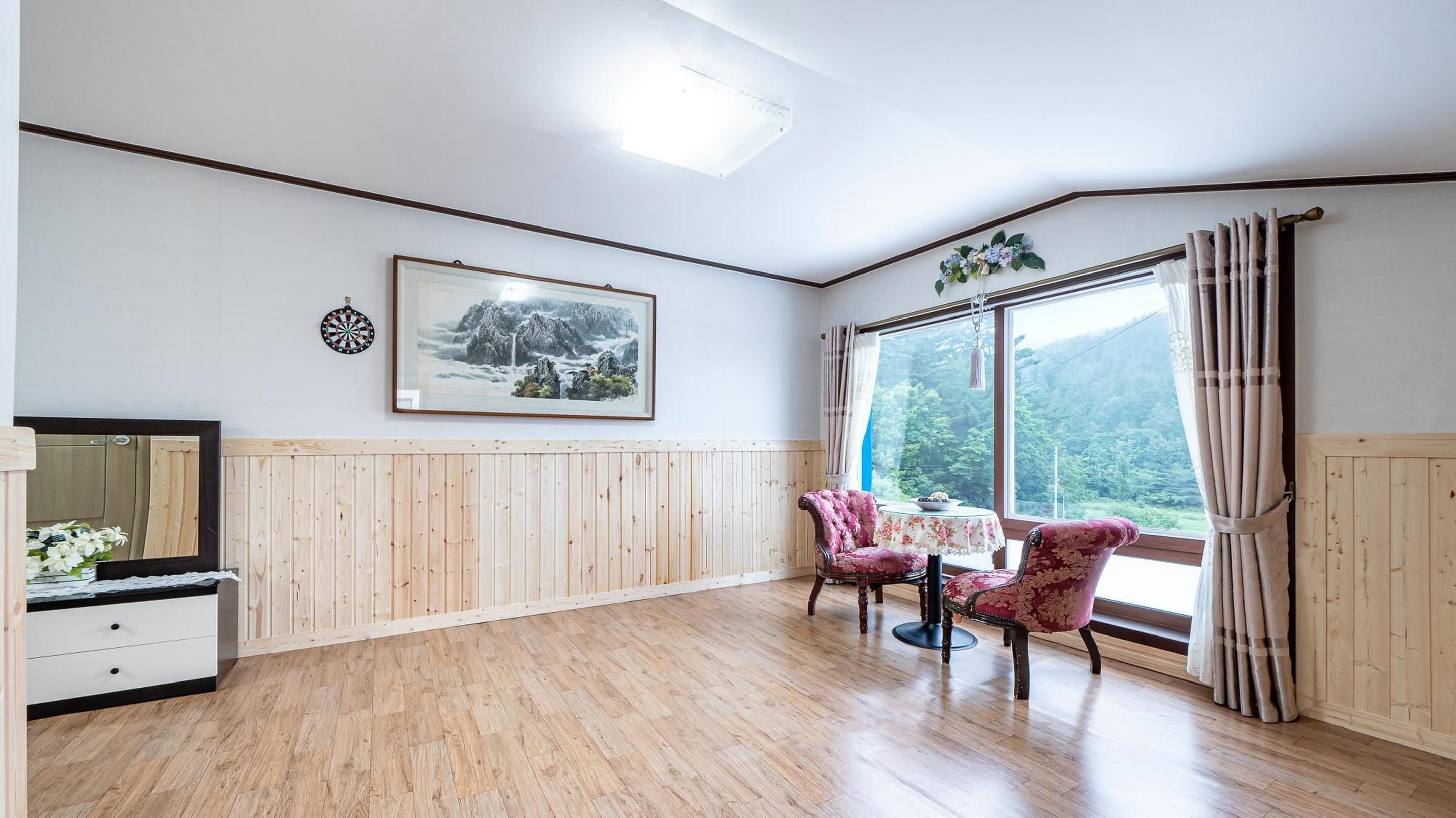 Una habitación con paneles de madera, un gran ventanal con cortinas, un cuadro de paisaje enmarco y dos sillas rosadas.