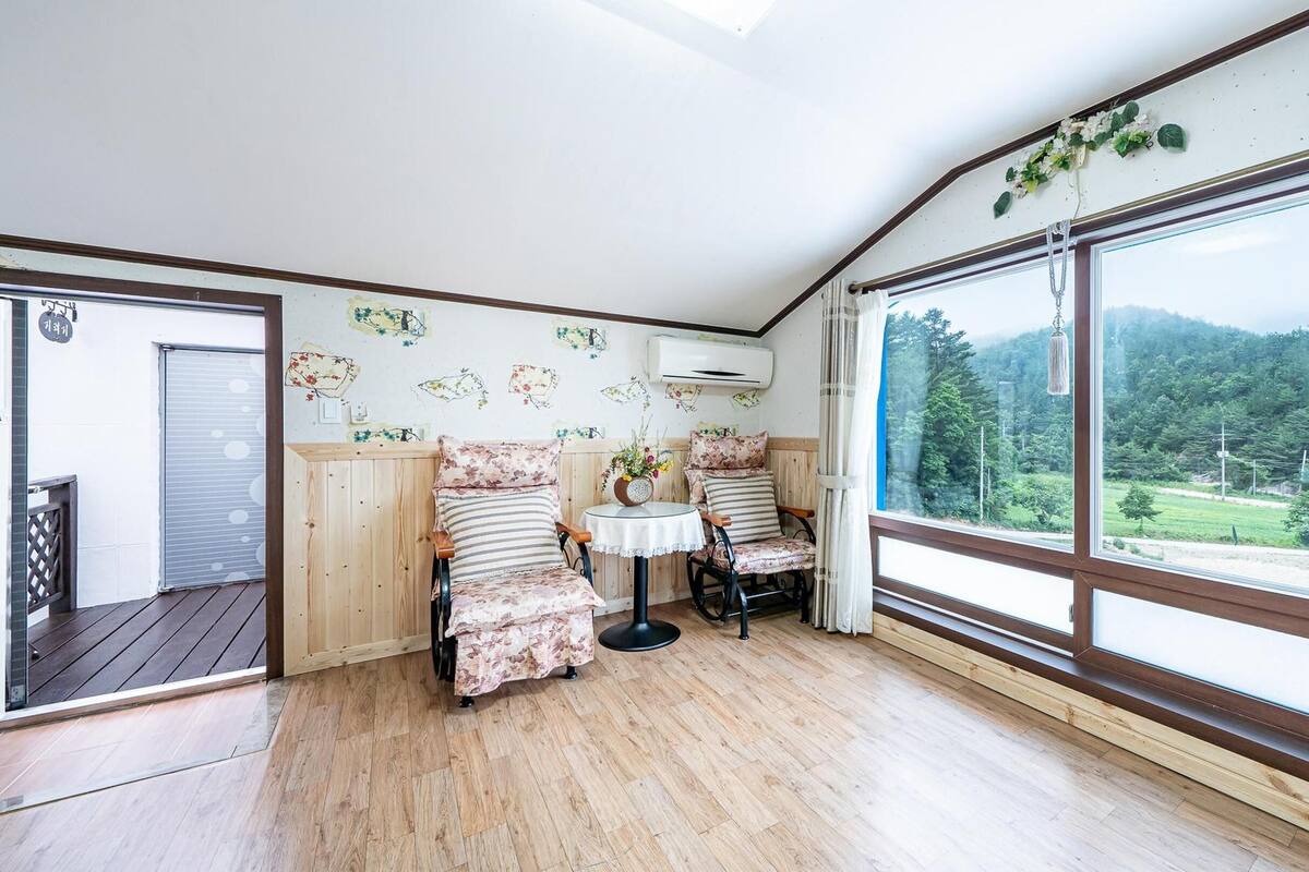 A cozy room with wooden flooring, two chairs with floral upholstery, a small round table, and a large window offering a view of a forested landscape.