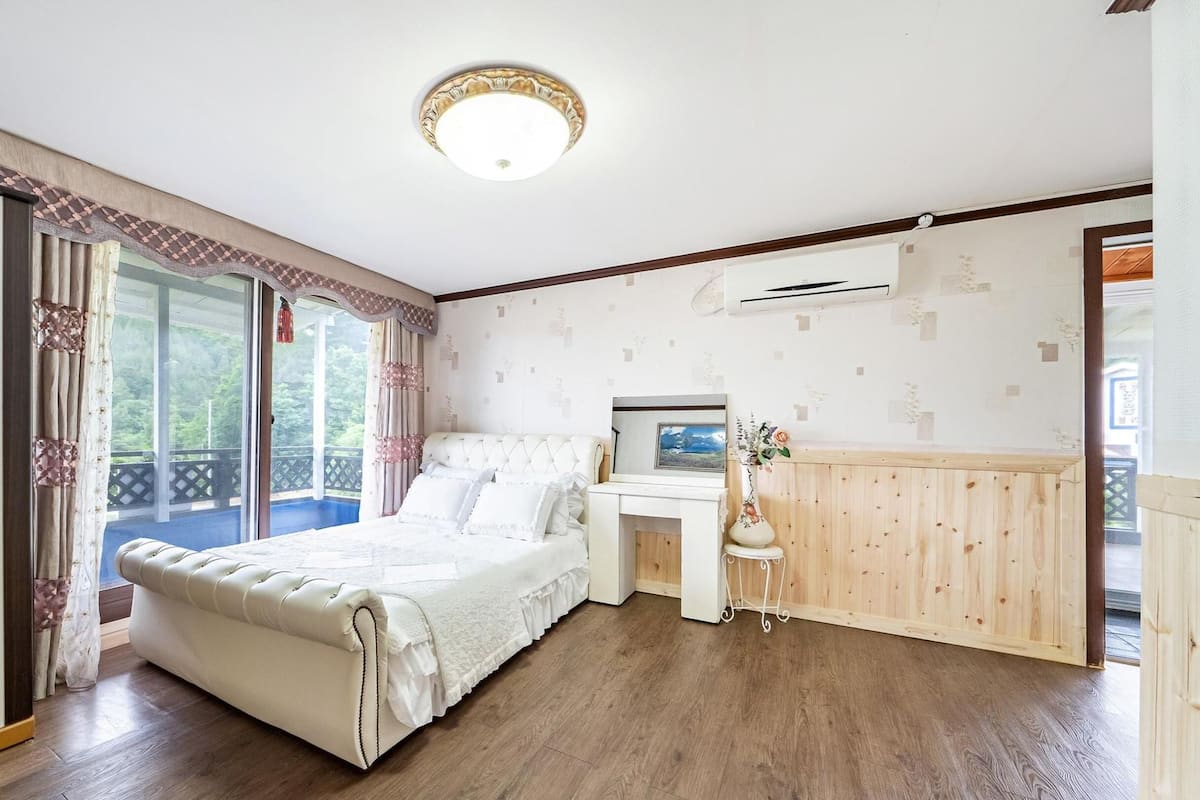 A bedroom with a large bed, a small table with a vase, and a view of the outdoors.
