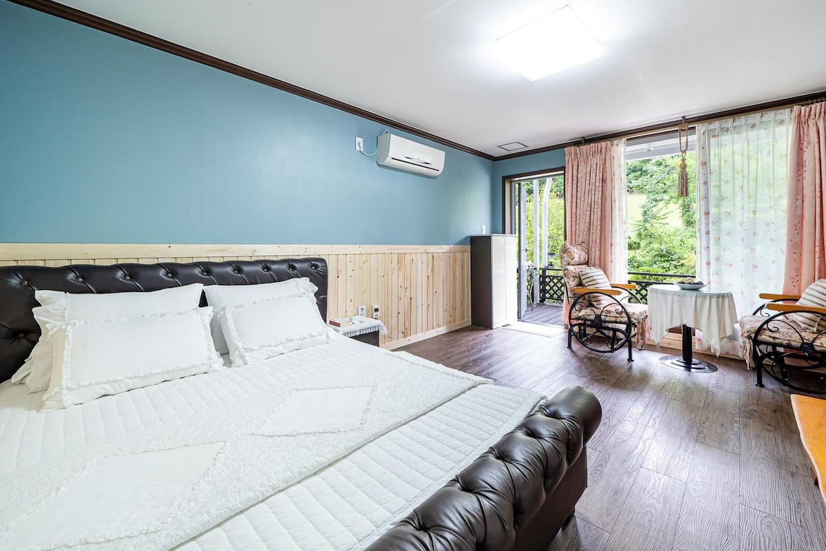 A bedroom with a bed, a wooden headboard, a nightstand, a window with curtains, and a chair by the window.