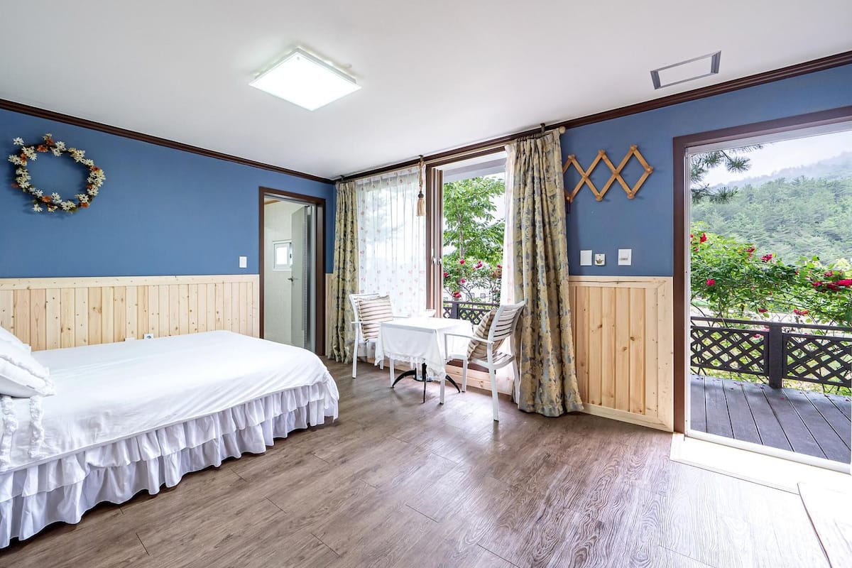 A bedroom with a wooden bed, a small table with chairs, a view of greenery through a large window, and a decorative wreath on the wall.