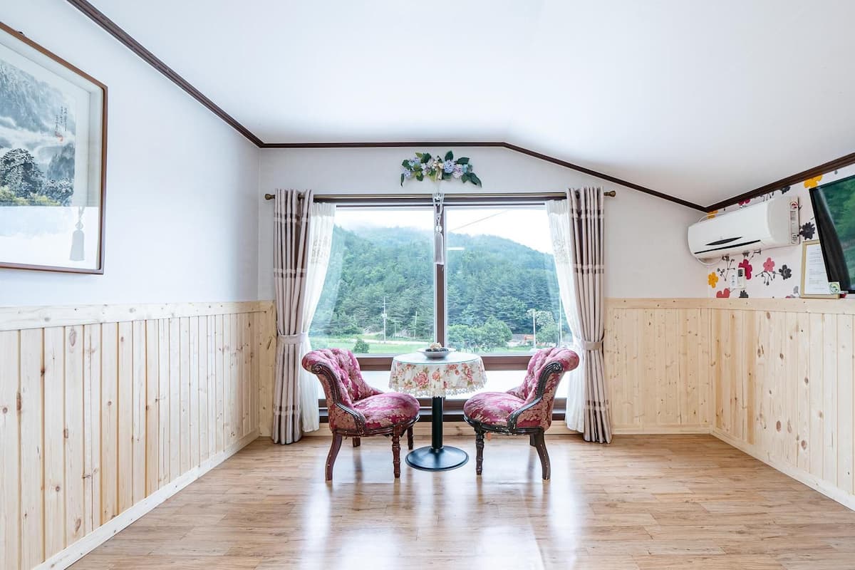 A room with wooden paneling, a table with chairs, a TV, and a window with a view of a forest.