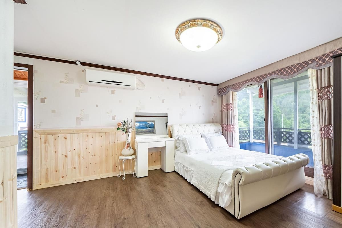 A bedroom with a large bed, a small bedside table, a wall-mounted air conditioner, and a window with curtains.