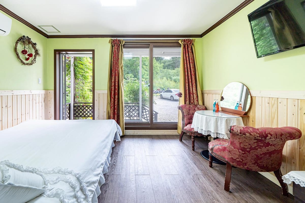 A room with a bed, a table with chairs, a mirror, and a view of the outside.