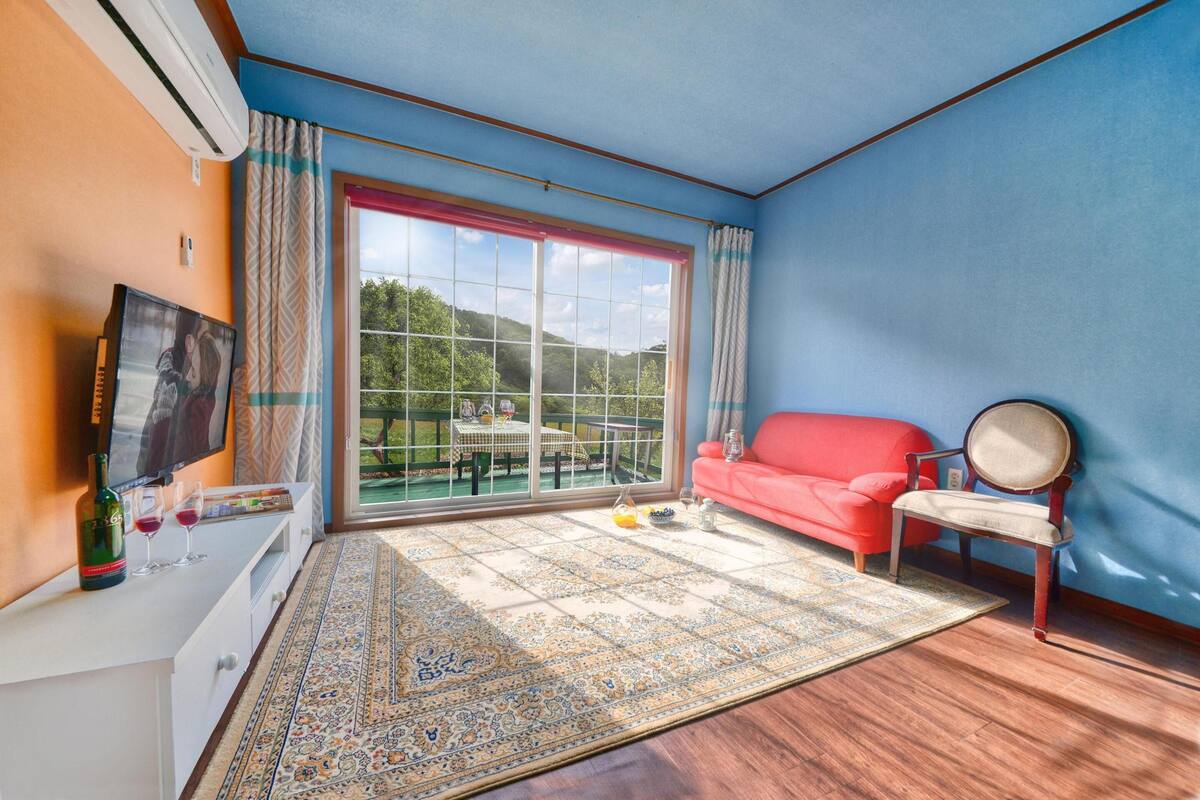 A room with a large sliding glass door to a balcony, a red sofa, a wooden chair, a TV, and a small table with a bottle and glasses.