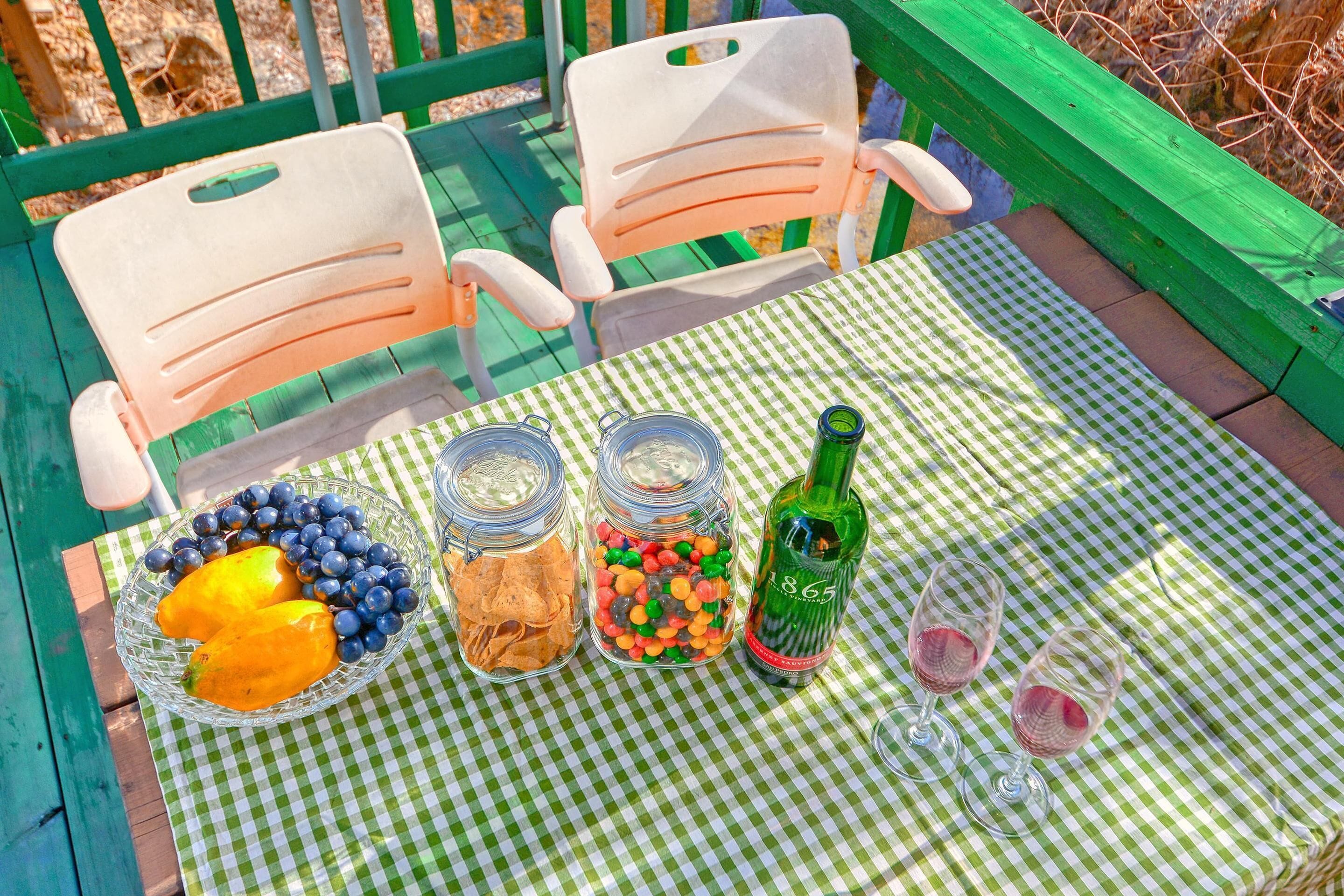 A picnic setup with a checkered tablecloth, two chairs, a bowl of fruit, jars of snacks, a bottle of wine, and two wine glasses.