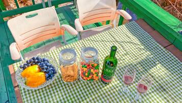A picnic setup with a checkered tablecloth, two chairs, a bowl of fruit, jars of snacks, a bottle of wine, and two wine glasses.