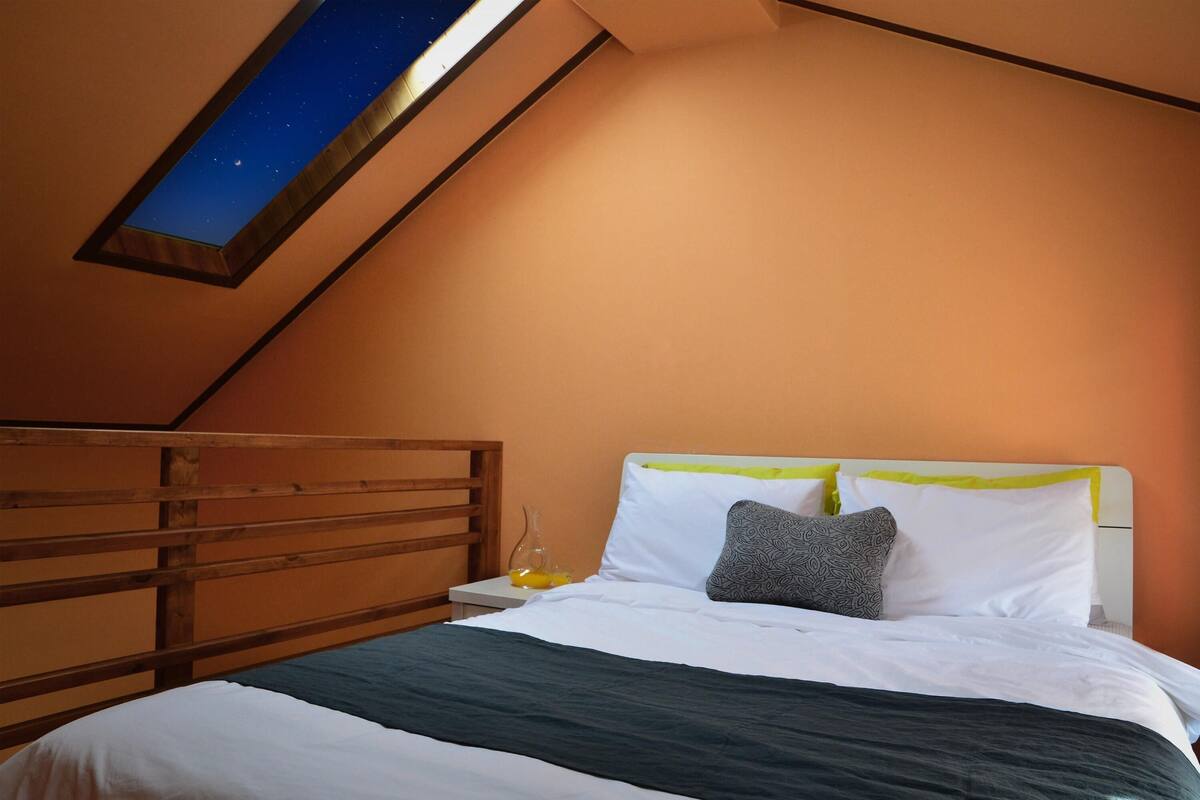 A bedroom with a sloped ceiling, a bed with white and green bedding, a wooden headboard, and a night sky view through a skylight.