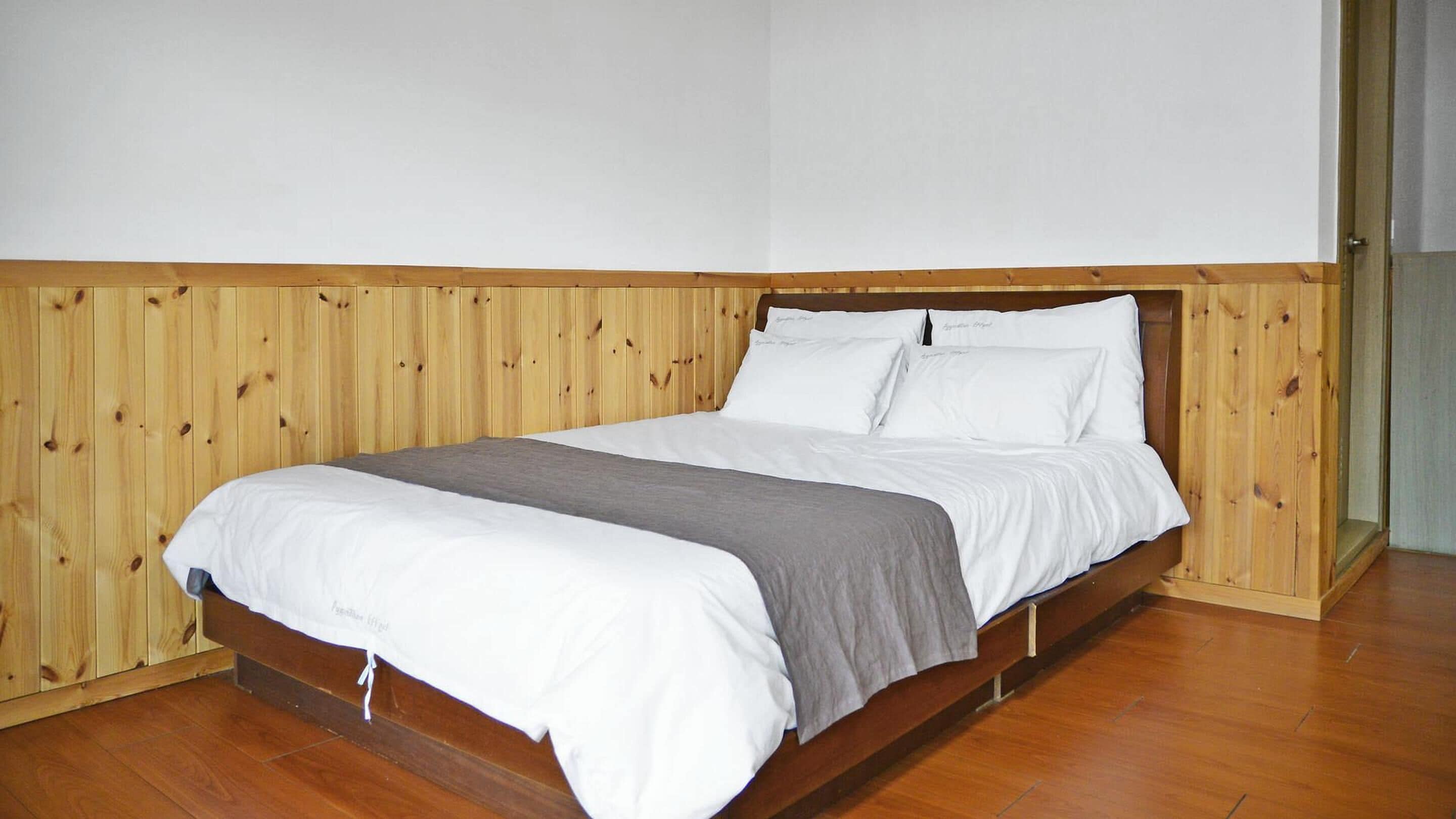 Una cama individual con cabecera de madera y una manta gris, sobre un suelo y pared de madera.