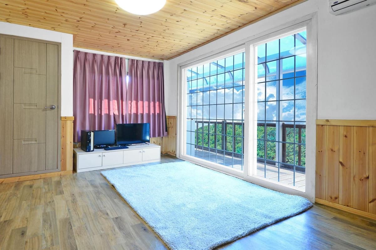 A room with a wooden ceiling, a large window with a view, a TV on a white cabinet, and a wooden wardrobe.