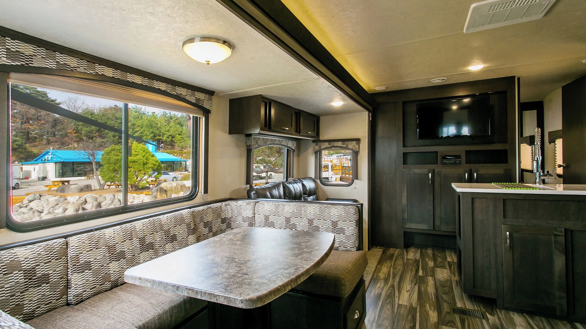 A compact RV kitchen with a dining area, a large window, and a built-in TV.