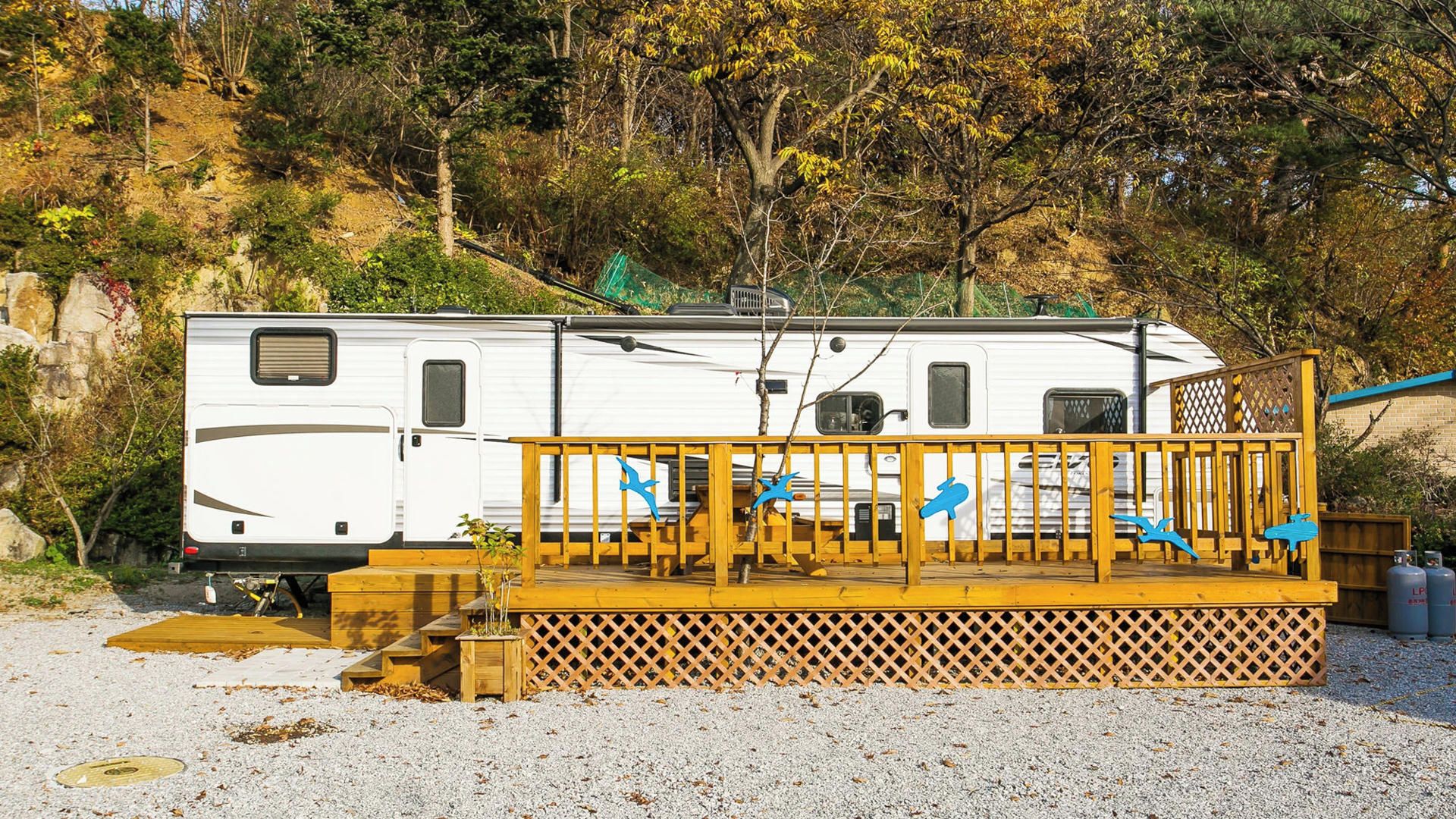 Een witte camper op grind met een houten terras en blauwe vogelbeeldjes.
