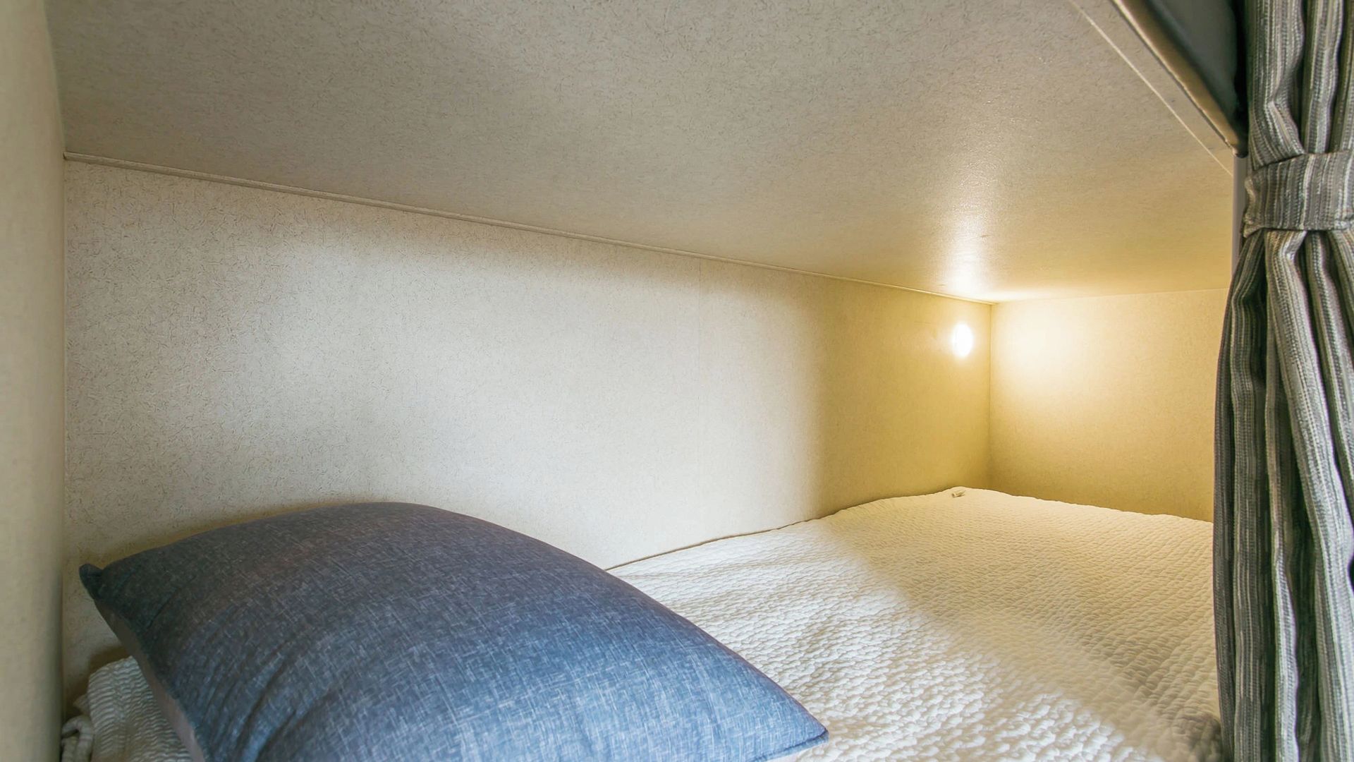 Una cama individual con un almohadón y una lámpara de pared en una habitación con paredes lisas.