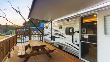 A covered deck with picnic tables, a white RV with a black design, and a blue building in the background.