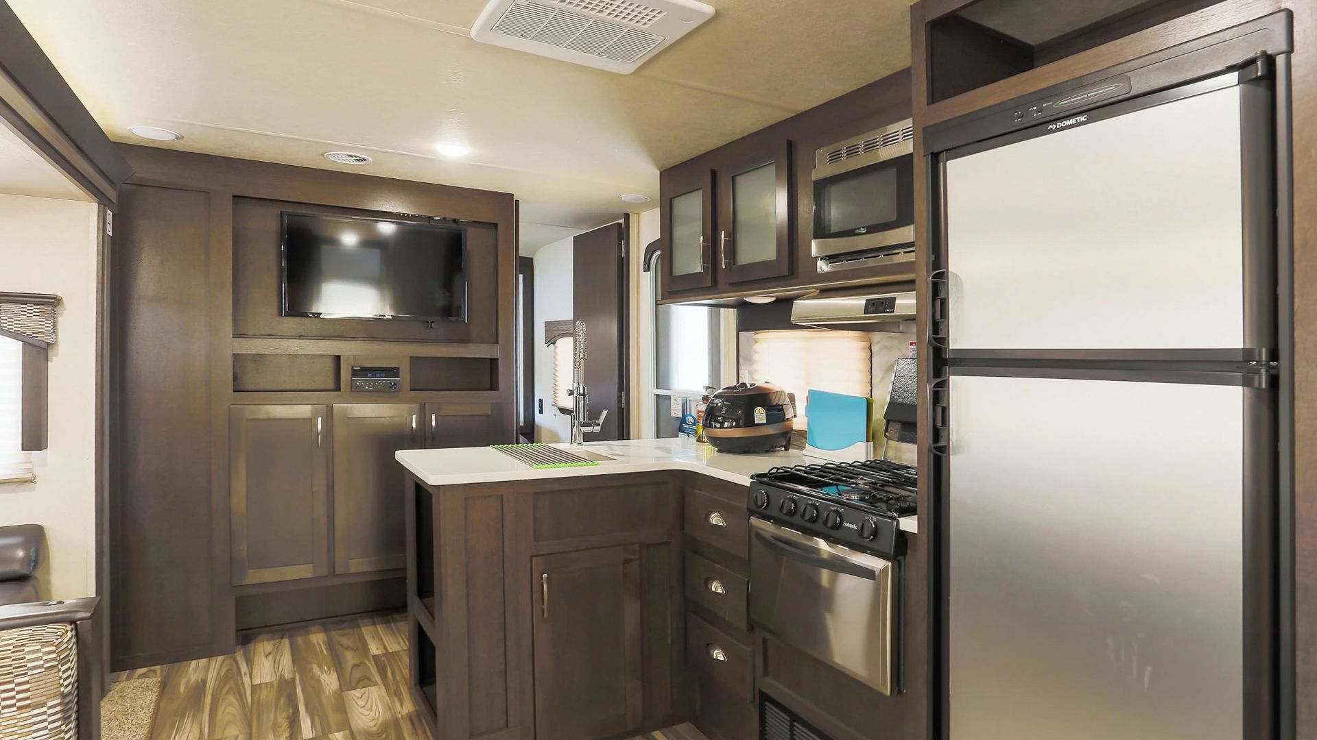 A compact RV kitchen with a stove, microwave, refrigerator, and a flat-screen TV.
