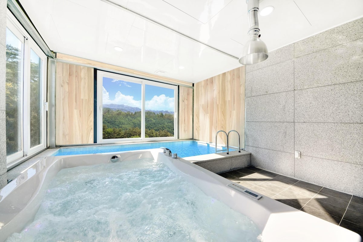 A modern bathroom with a large bathtub, a small pool, and a view of a mountainous landscape through large windows.