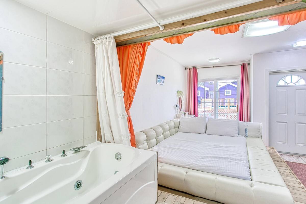 A bedroom with a large bed, a bathtub, and a view of a blue house through the window.
