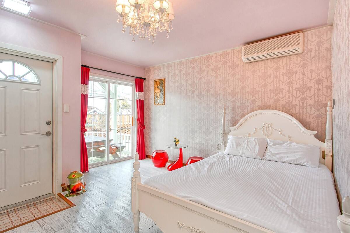 A bedroom with a white bed, a chandelier, a red table, and a window with curtains.