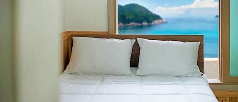 A bed with white bedding and a wooden headboard, positioned next to a window with a view of a coastal landscape.